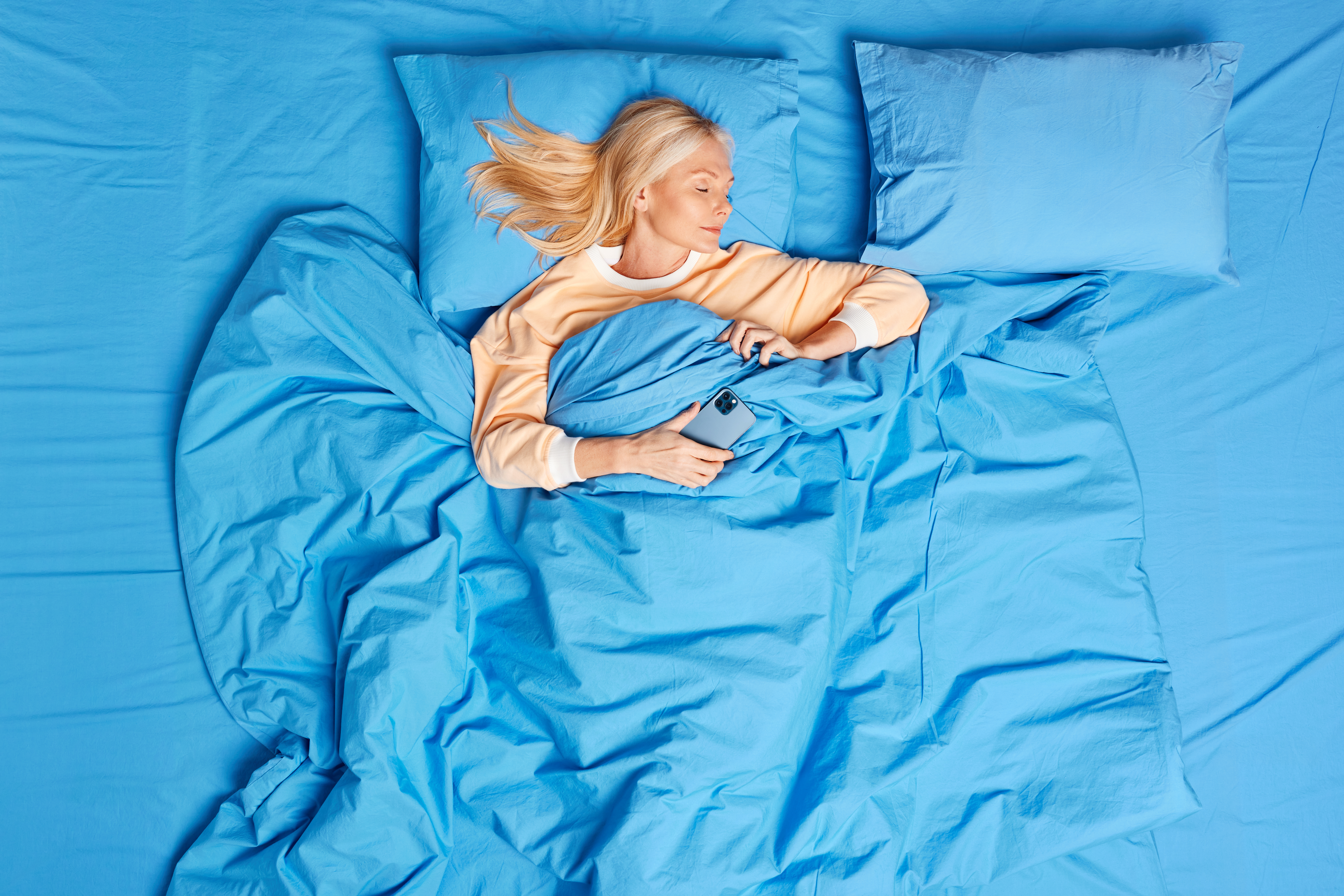 Beautiful middle aged blonde woman has technology addiction sleeps at comfortabled bed with smartphone takes nap and waits for important call being always in touch. Rest and technology concept